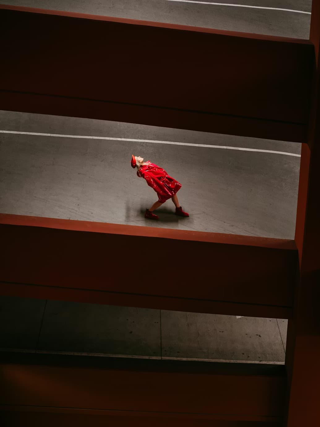 Person in red leans back dramatically in a parking garage framed by dark horizontal beams.